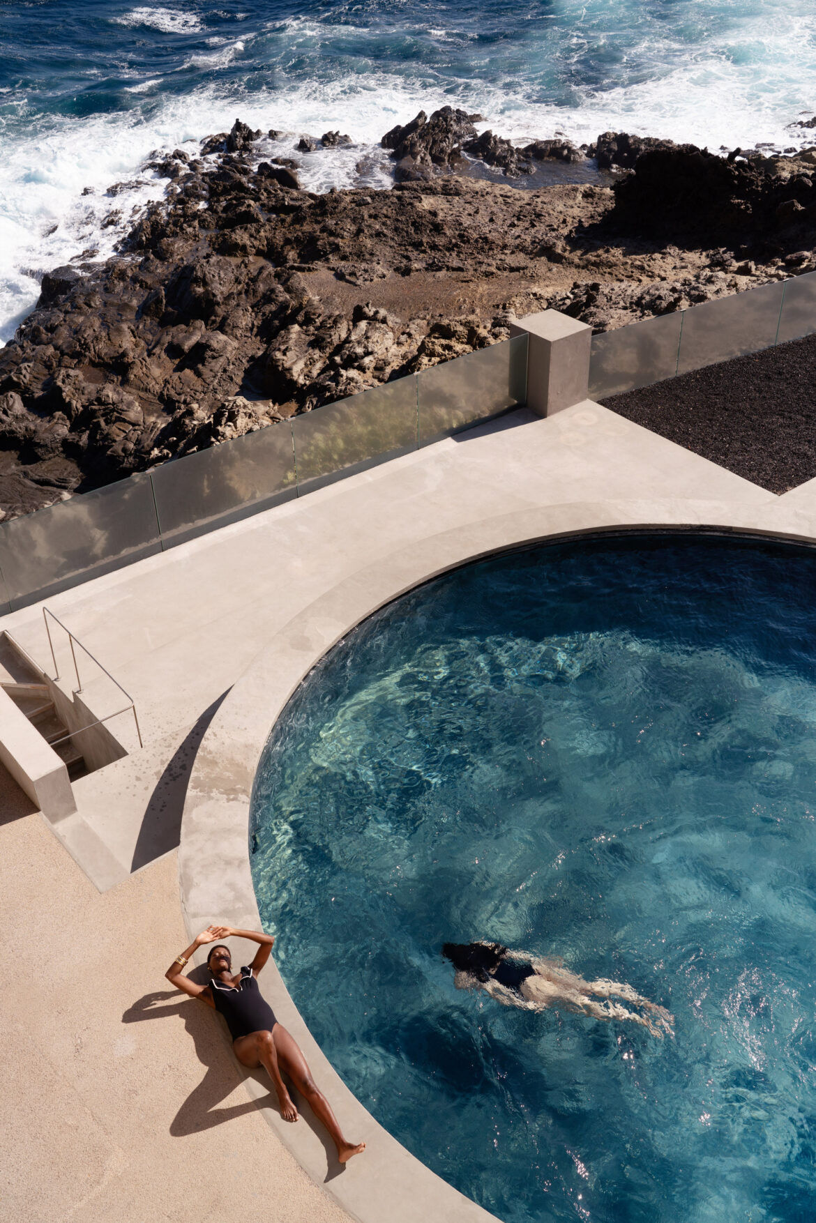 Fashion Photographer London Fashion-forward editorial photography by Pedro Antunes featuring a serene poolside scene: one model floats gracefully in a round, crystal-clear pool while another in a chic black swimsuit lounges under the sun, evoking luxury and relaxation. Surrounded by dramatic rocky cliffs and the sparkling ocean beyond, this high-end fashion campaign image blends natural textures with sophisticated styling—perfect for e-commerce, social media content, and brand storytelling that captures elegance, summer lifestyle, and modern aesthetics.