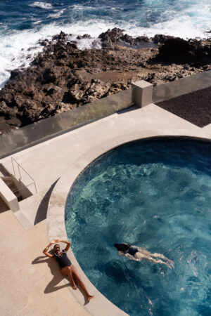 Fashion Photographer London Fashion-forward editorial photography by Pedro Antunes featuring a serene poolside scene: one model floats gracefully in a round, crystal-clear pool while another in a chic black swimsuit lounges under the sun, evoking luxury and relaxation. Surrounded by dramatic rocky cliffs and the sparkling ocean beyond, this high-end fashion campaign image blends natural textures with sophisticated styling—perfect for e-commerce, social media content, and brand storytelling that captures elegance, summer lifestyle, and modern aesthetics.