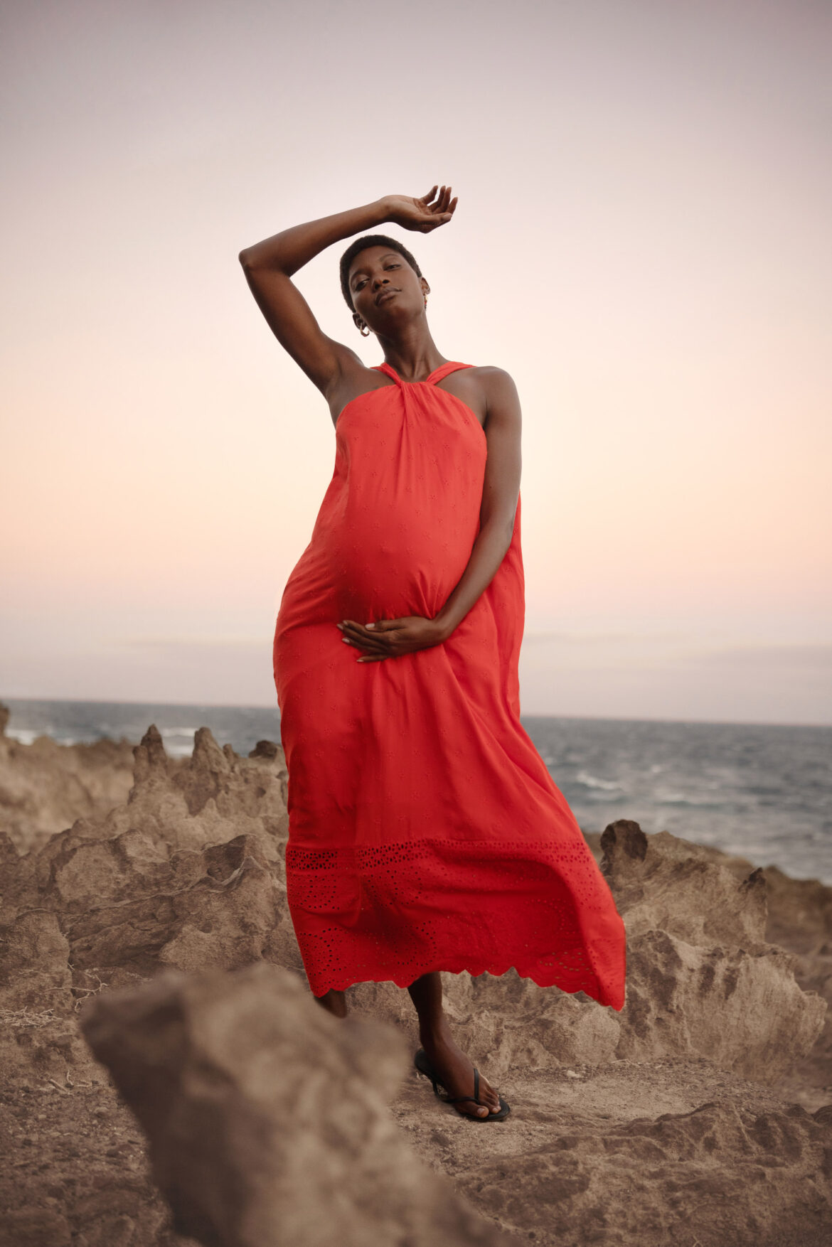 Fashion Photographer London A woman in a flowing red dress stands on rocky terrain by the sea at sunset, gently holding her baby bump with one hand and raising the other above her head. The sky is soft pastel hues, creating a serene atmosphere.